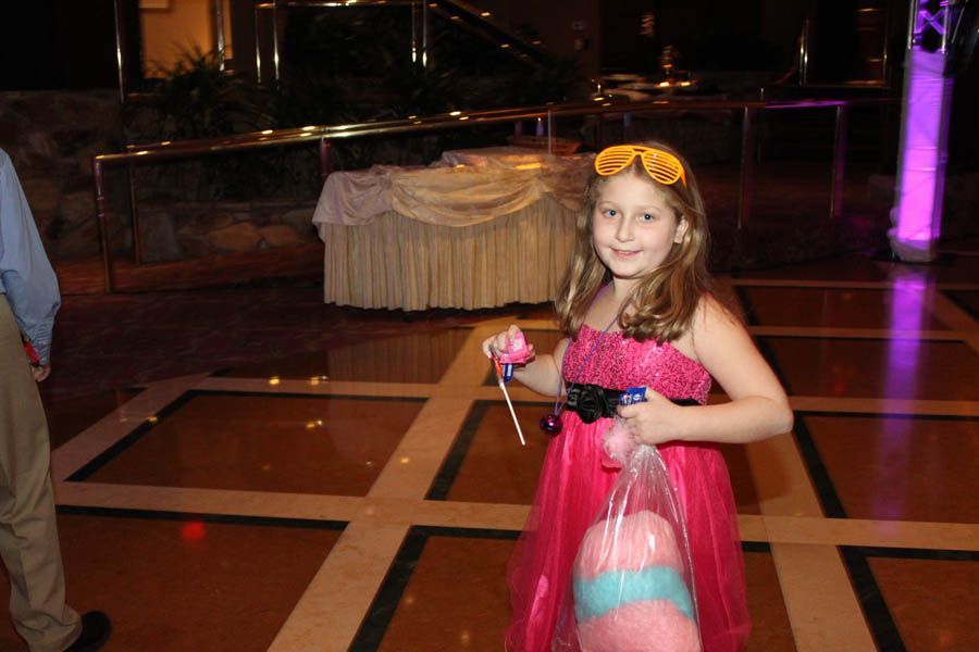 Girl in pink dress smiles, holding candy and cotton candy on a dance floor with a formal buffet setting.
