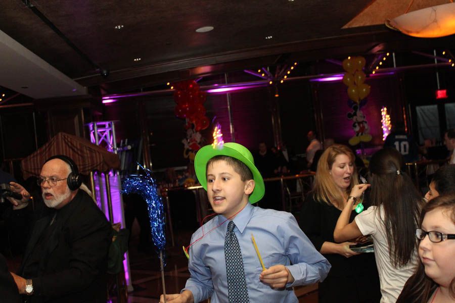 A young person in a green hat dancing at a party, surrounded by other guests and balloons.