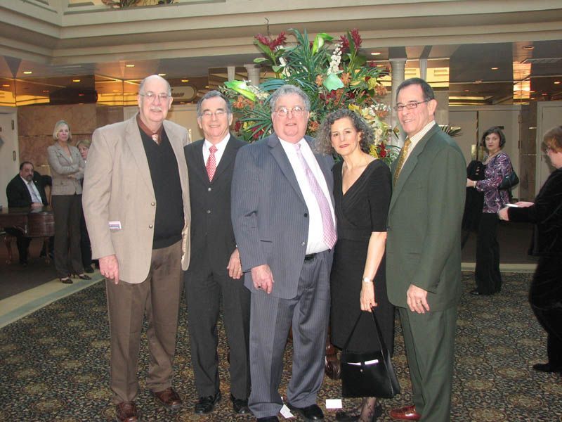 Five people pose near a floral arrangement indoors; carpeted floor, neutral colors.