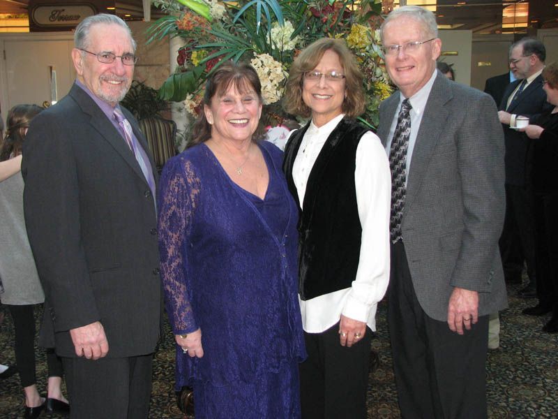 Four people smiling, posing in a well-lit space in front of floral arrangement. Formal attire.