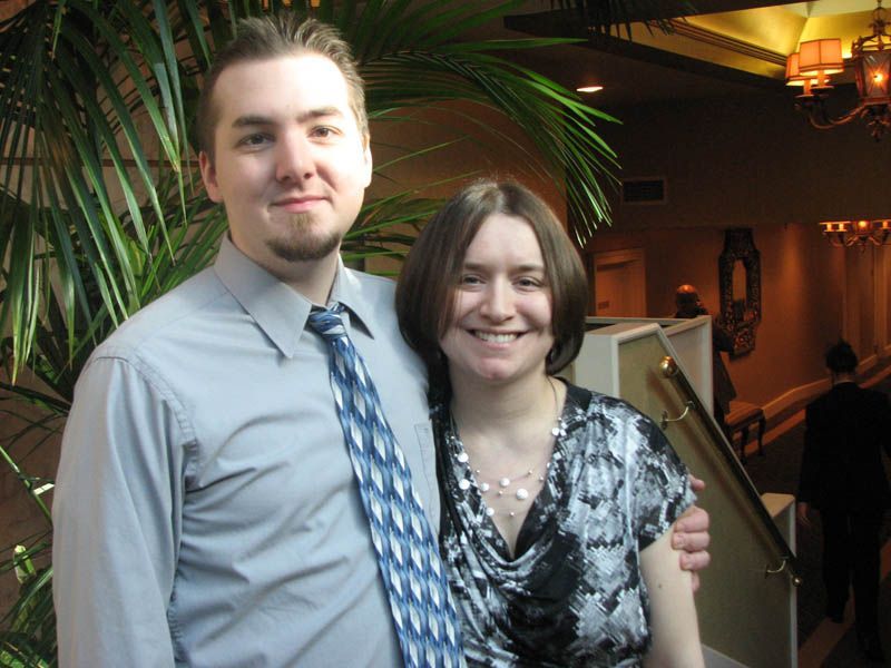 Man in gray shirt and tie with woman in patterned top, posing indoors.
