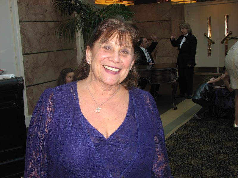 Woman smiles, wearing a purple lace top, standing indoors. People are in the background.