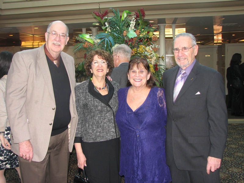Four people smiling, posing near a large flower arrangement. One woman wears purple, others in formal attire. Indoors.