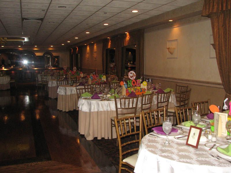 Long ballroom with tables set for a party, decorated with colorful centerpieces, and gold chairs.
