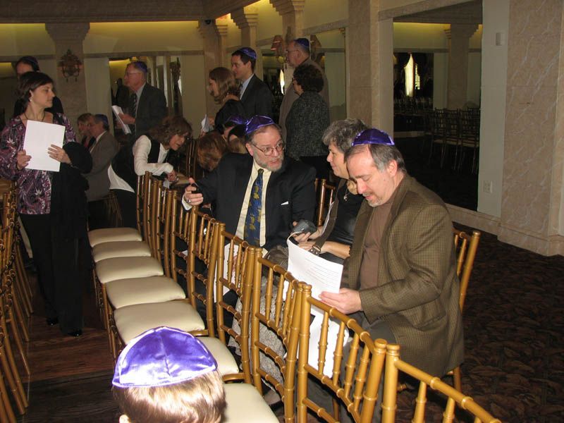 People wearing yarmulkes, some reading papers, in a decorated room with gold chairs.