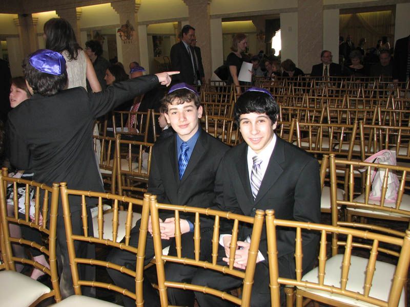 Two boys in suits and yarmulkes seated in a row of gold chairs; person points.