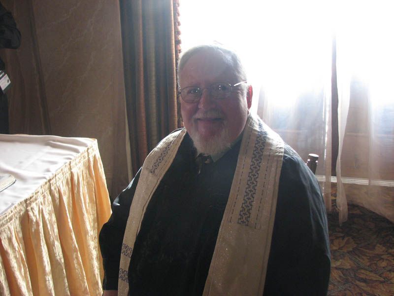 Man wearing glasses and a religious shawl, smiling, seated indoors near a window.