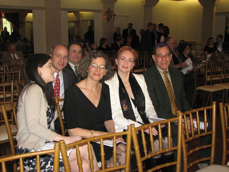 People seated in gold chairs at an event, some wearing suits and dresses, in a room with columns.