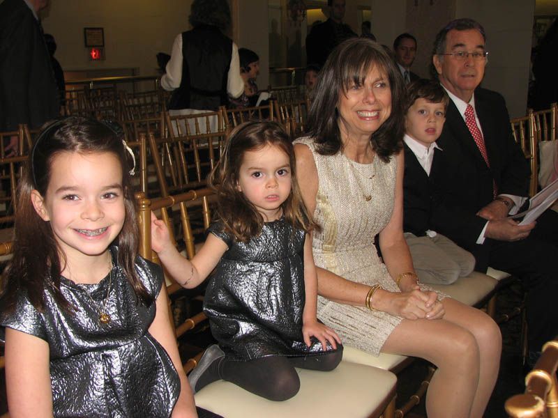 Family sitting in chairs at an event; girls in metallic dresses, woman in ivory dress, and man in suit.