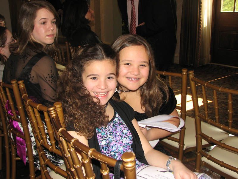 Two girls smiling, sitting in gold chairs. One has curly brown hair, the other has straight hair. Indoors, festive setting.
