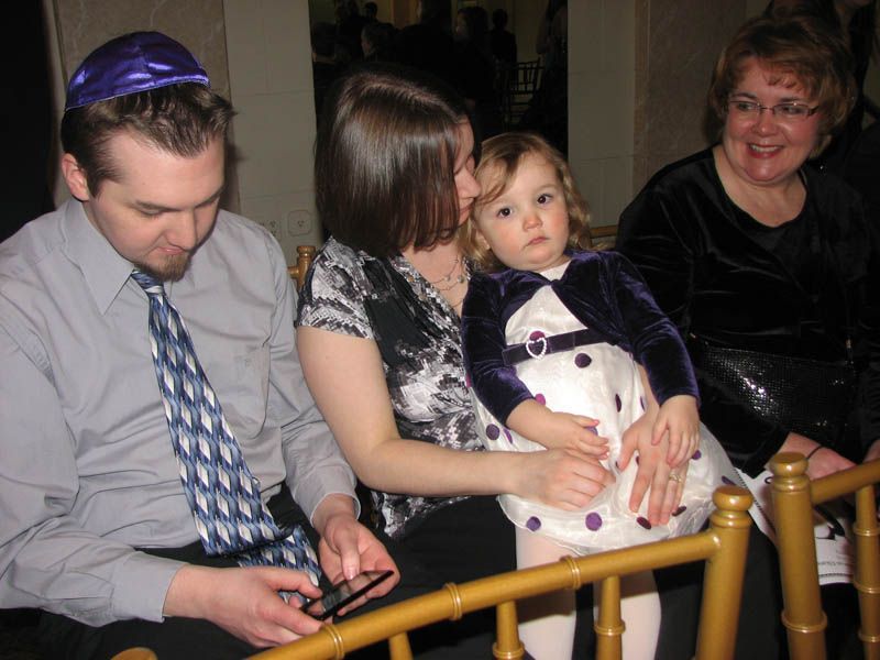 Family gathered; man with yarmulke, woman holding child, another woman smiles. Indoor setting, gold chairs.