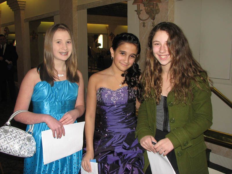Three smiling girls in formal dresses indoors, holding papers.