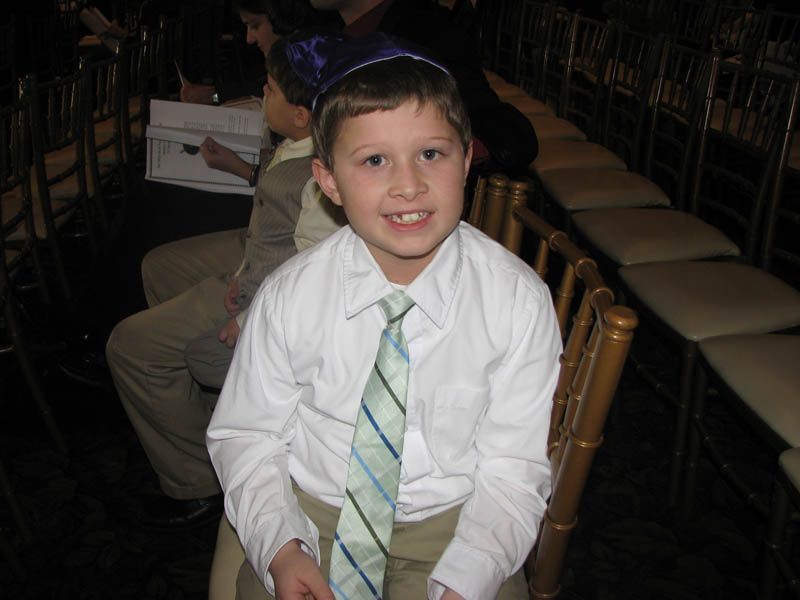 Boy wearing a kippah and tie smiles, sitting in a chair at an event.