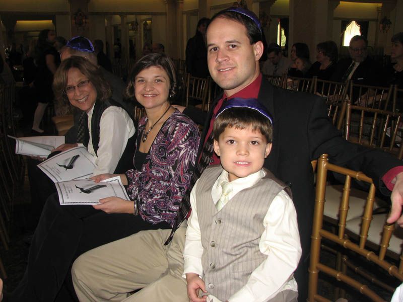 Family seated in chairs, all wearing yarmulkes. The son is in a vest and the father has a suit jacket.