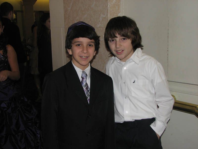 Two boys in formal attire smiling near a wall. One in a suit, the other in a button-down shirt.