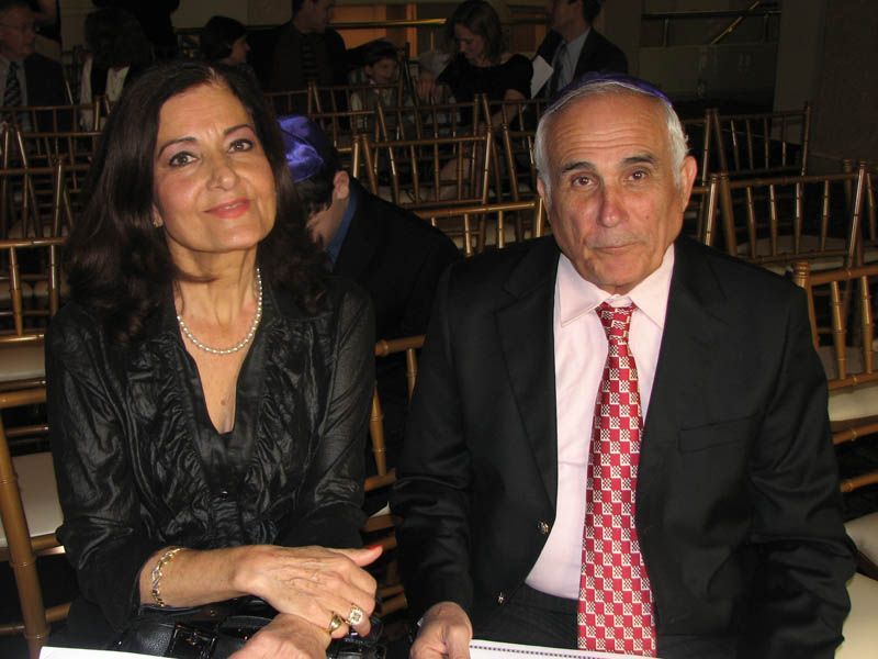 Woman and man seated, smiling, in formal attire; gold chairs in background.