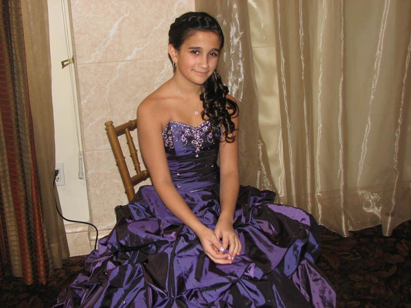 Young person in a purple formal gown sits on a chair near curtains, smiling.