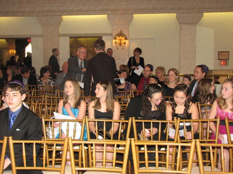 People seated in ornate chairs at an event; hall with columns. Some wear formal attire; several looking at something.
