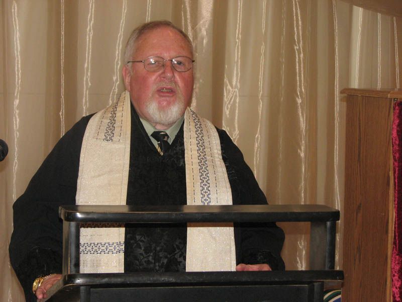 Man speaking from a podium, wearing a black robe and a white shawl, beige curtain backdrop.
