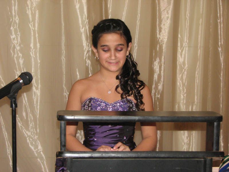Woman in a purple dress, at a podium, with microphone; curtain backdrop.