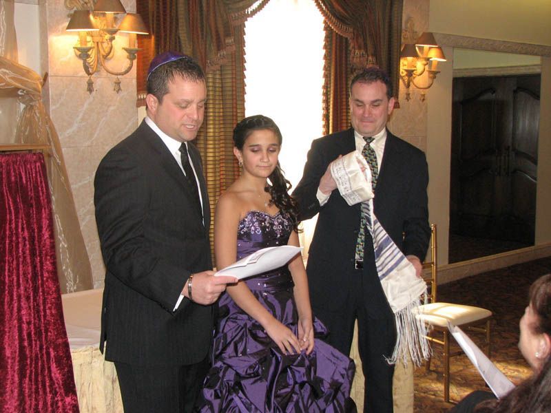 People at a Bar Mitzvah: a young person in a purple dress with two men, one holding a tallit, inside a room.