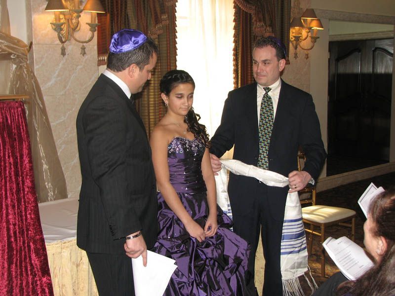 Two men place a prayer shawl on a young person wearing a purple dress, indoors.