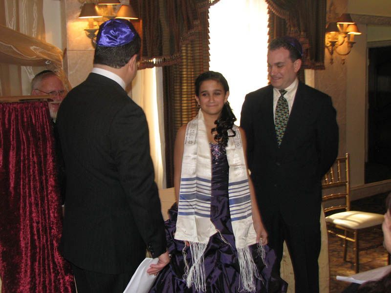 Girl in purple gown wearing a tallit, with two men, likely at a Bat Mitzvah ceremony.