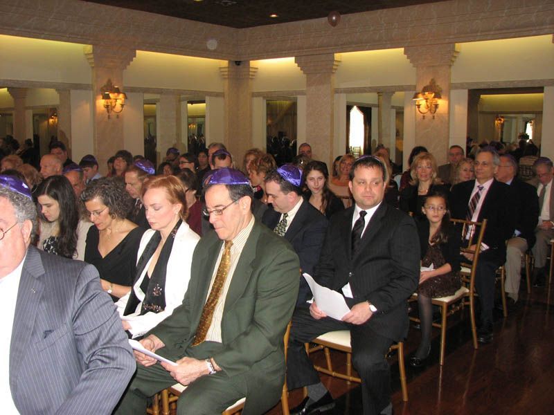 People seated at a ceremony, many wearing yarmulkes, in a well-lit hall with pillars and ornate light fixtures.