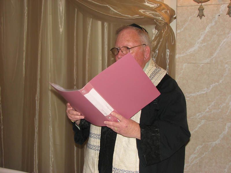 Man in black robe with tallit, reading from a pink folder. Beige curtain background.