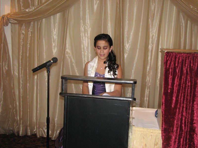 Young person in formal attire speaking at a podium, microphone present, stage curtains backdrop.