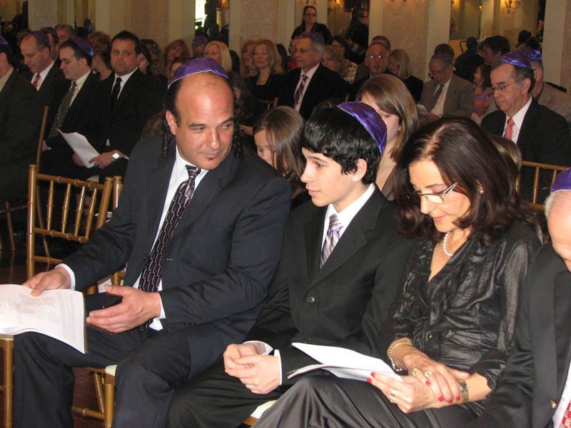 Family at an event; man, teen, woman seated, all with yarmulkes, reading papers. Other attendees in background.