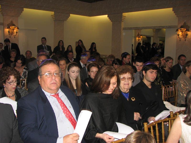 People gathered inside a room with ornate columns, likely a formal event. Many are wearing yarmulkes, some holding papers.