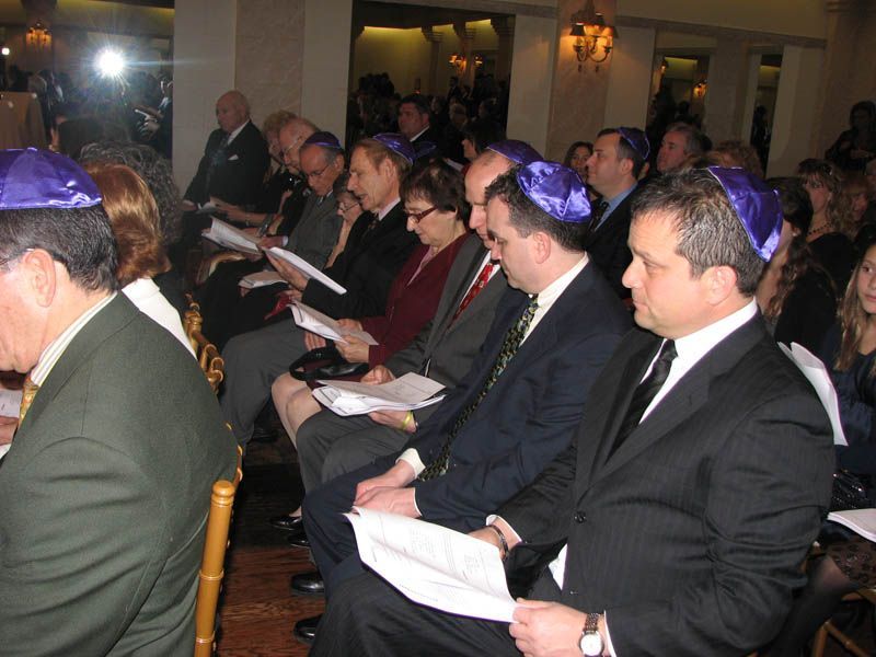 People seated, some wearing purple yarmulkes, reading papers indoors.
