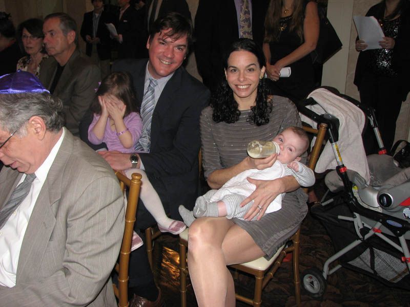 Family at an event; mother feeding baby, father and daughter seated. Other guests in the background.