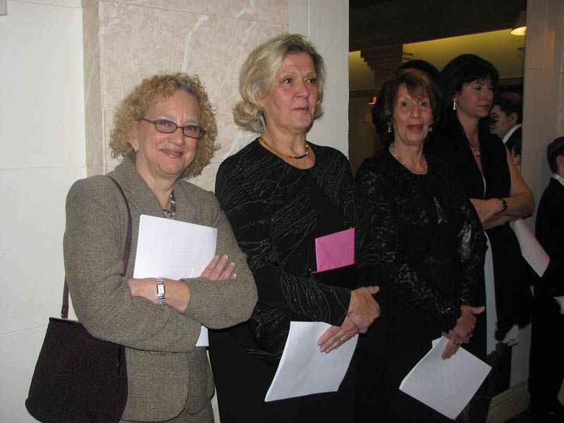 Three women standing, holding papers, in a formal setting. The woman on the left has crossed arms. Others in background.