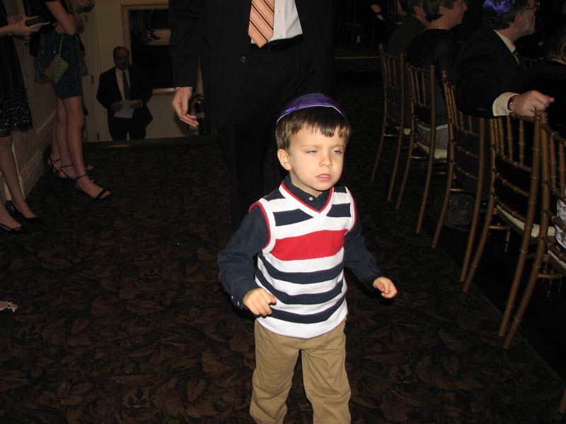 Boy in striped vest and yarmulke dancing on a dark carpet, another person in the background.