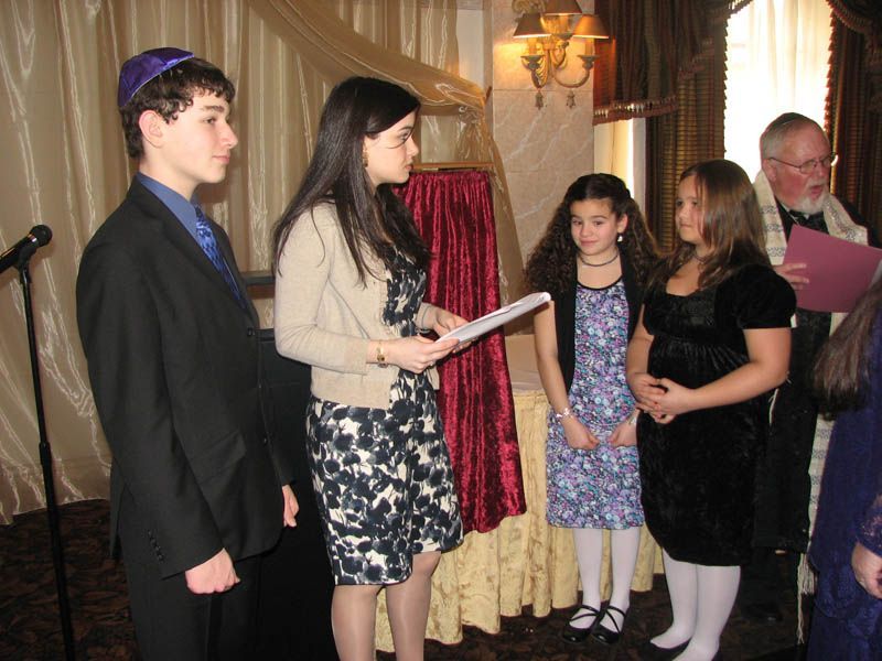 Group of people at a ceremony, reading papers. A person in a kippah. Indoor setting with curtains.