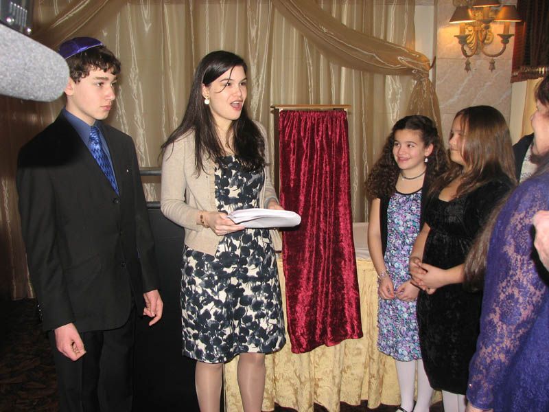 Woman speaking at an event, holding papers, with two girls and a boy in formal attire standing nearby.