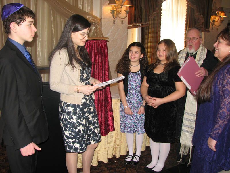 People at a Jewish ceremony. A woman reads a document; others stand by, wearing formal attire.