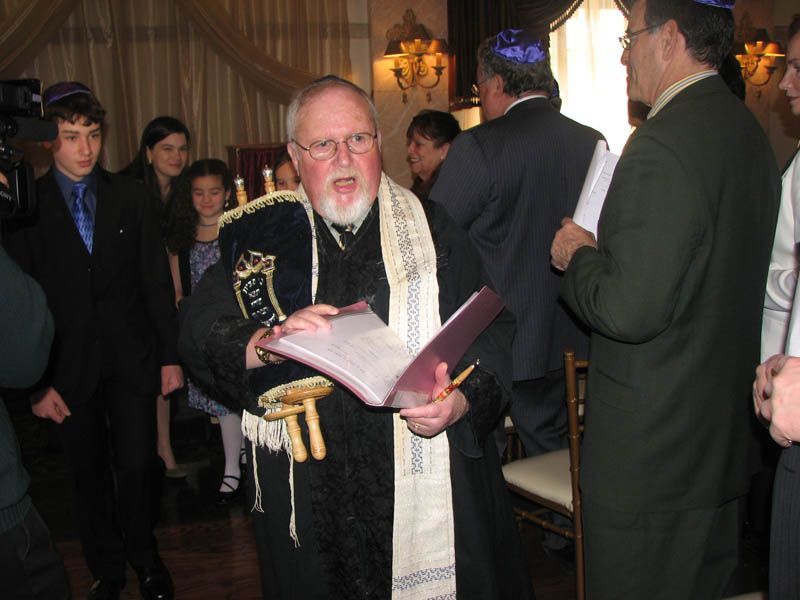 A man in religious attire reads from a book, possibly leading a ceremony. Indoors, others observe.