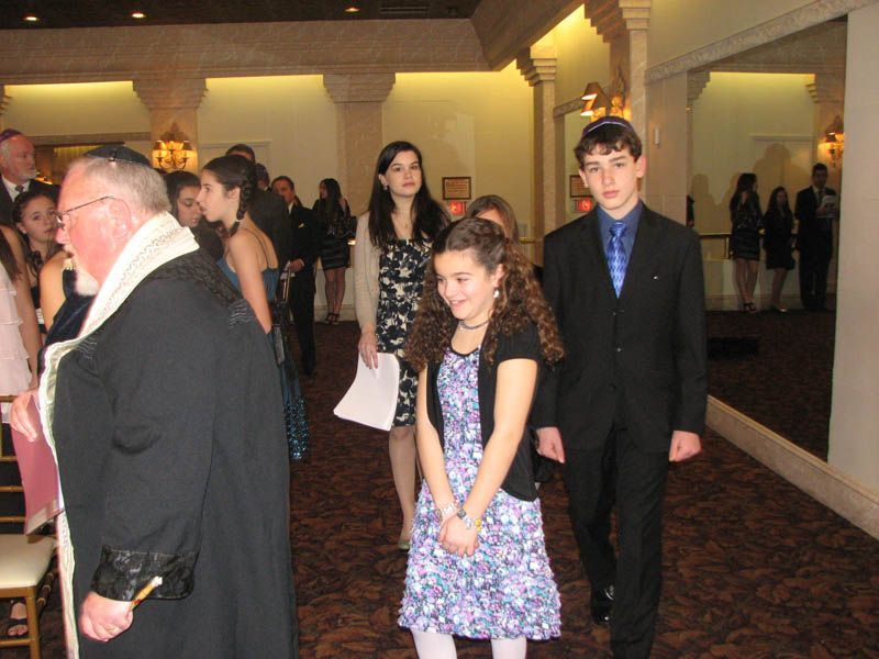 People in formal attire walking in a venue with ornate walls. A rabbi in traditional garb leads the procession.