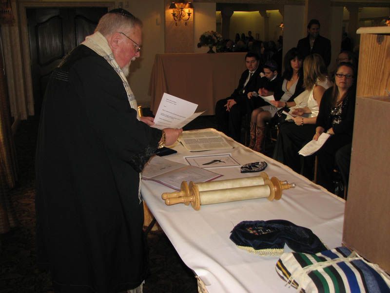 Rabbi reads from papers at a table with a Torah scroll; guests sit in background.