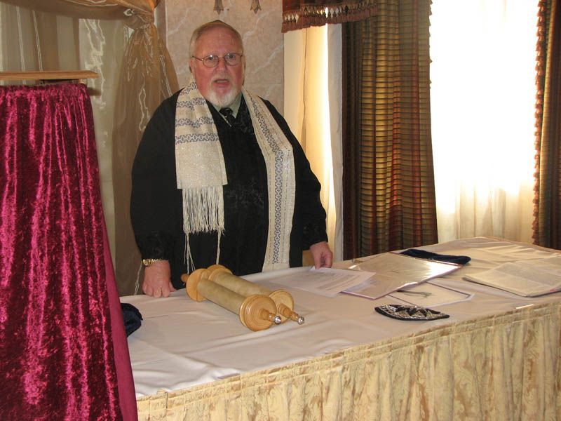 Man in black robe and tallit stands at table with Torah scroll, documents.