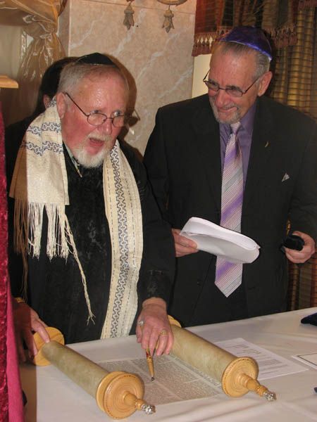 Two men reading a Torah scroll. One wears a tallit, the other a yarmulke and glasses, both smiling.