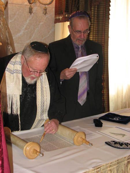 Two men reading a Torah scroll at a table; one wears a tallit and kippah, the other a kippah and suit.