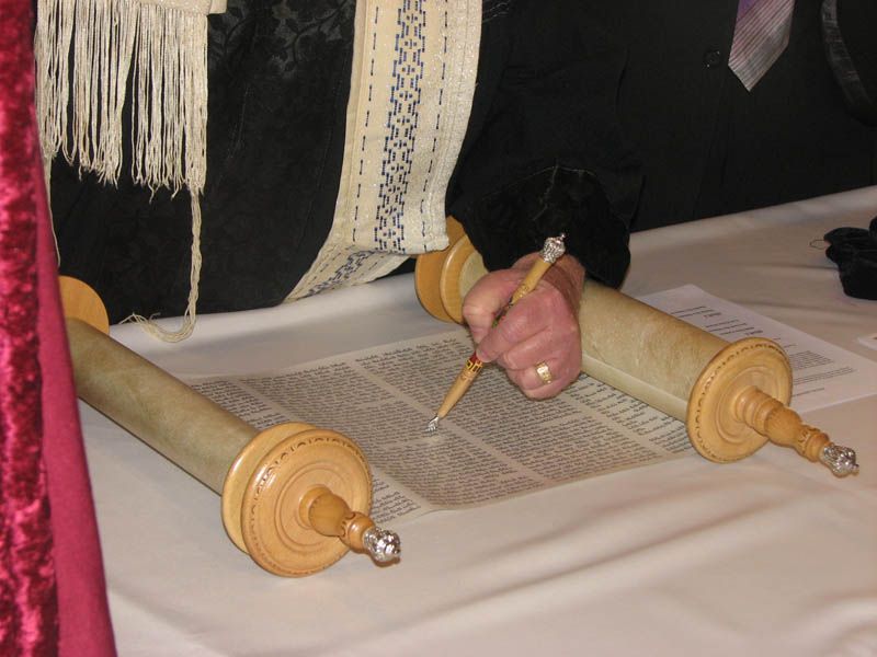 Person writing with a quill on a Torah scroll, wearing a tallit.