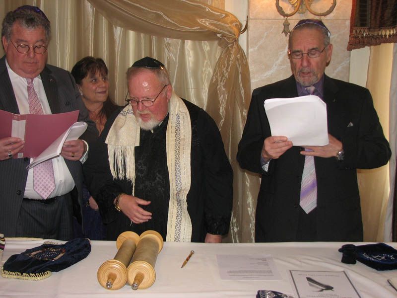 Men in suits and Jewish prayer shawls read from papers at a table with Torah scrolls, indoors.