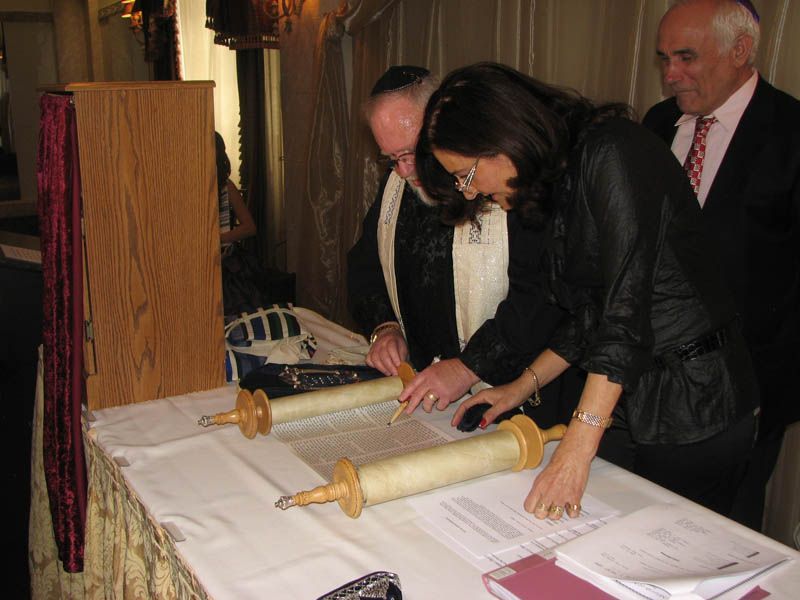 People signing a document near a Torah scroll on a table in a formal setting.