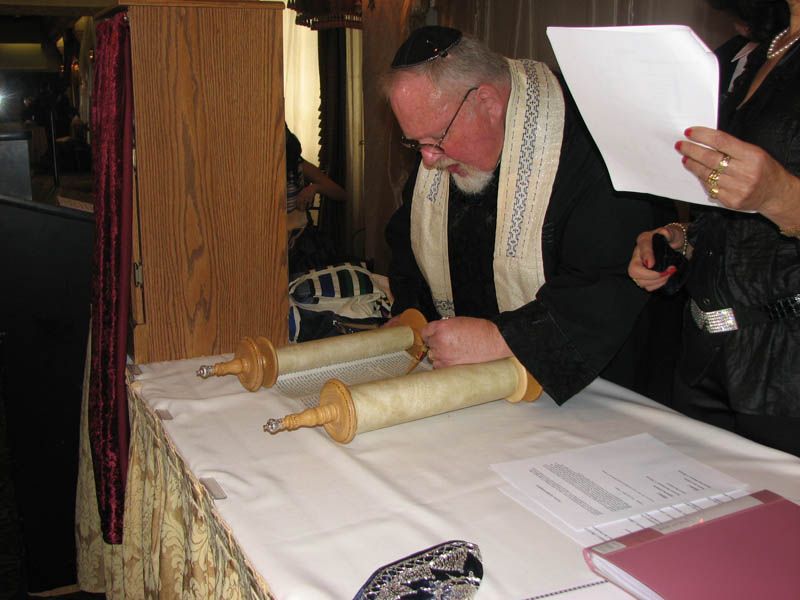 A rabbi wearing a kippah and tallit, scrolls a Torah at a table in a room.