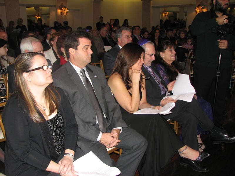 People seated at an event; indoor setting. Some are looking forward, others looking down, holding papers.